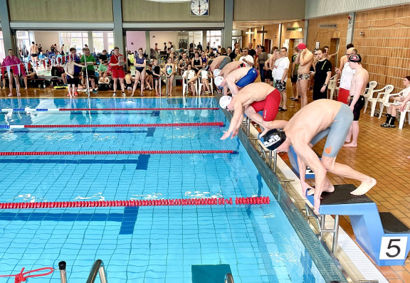 Dutzende Zuschauer stehen um ein Schwimmbecken herum, von dessen Startblöcken gerade fünf sportliche Jungen in Badekleidung und mit Badekappen springen.