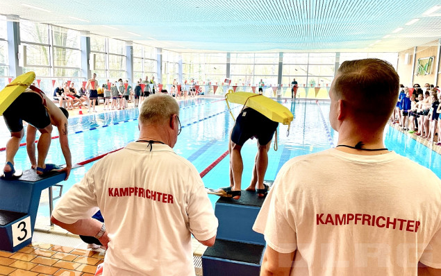 Zwei Kampfrichter mit entsprechendem Aufdruck auf dem Shirt stehen mit dem Rücken zur Kamera. Sie beobachten zwei Sportler mit je einem Gurtretter auf dem Rücken, die auf den Startblöcken eines Schwimmbeckens stehen.